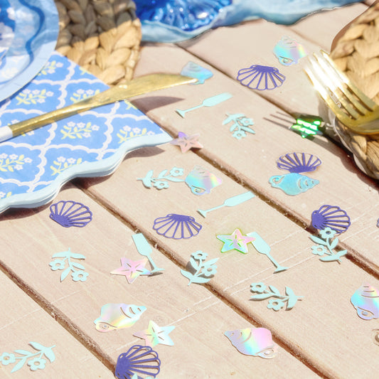 Beach themed bridal shower table styled with blue plates, gold flatware and coastal confetti with seashell and starfish designs.