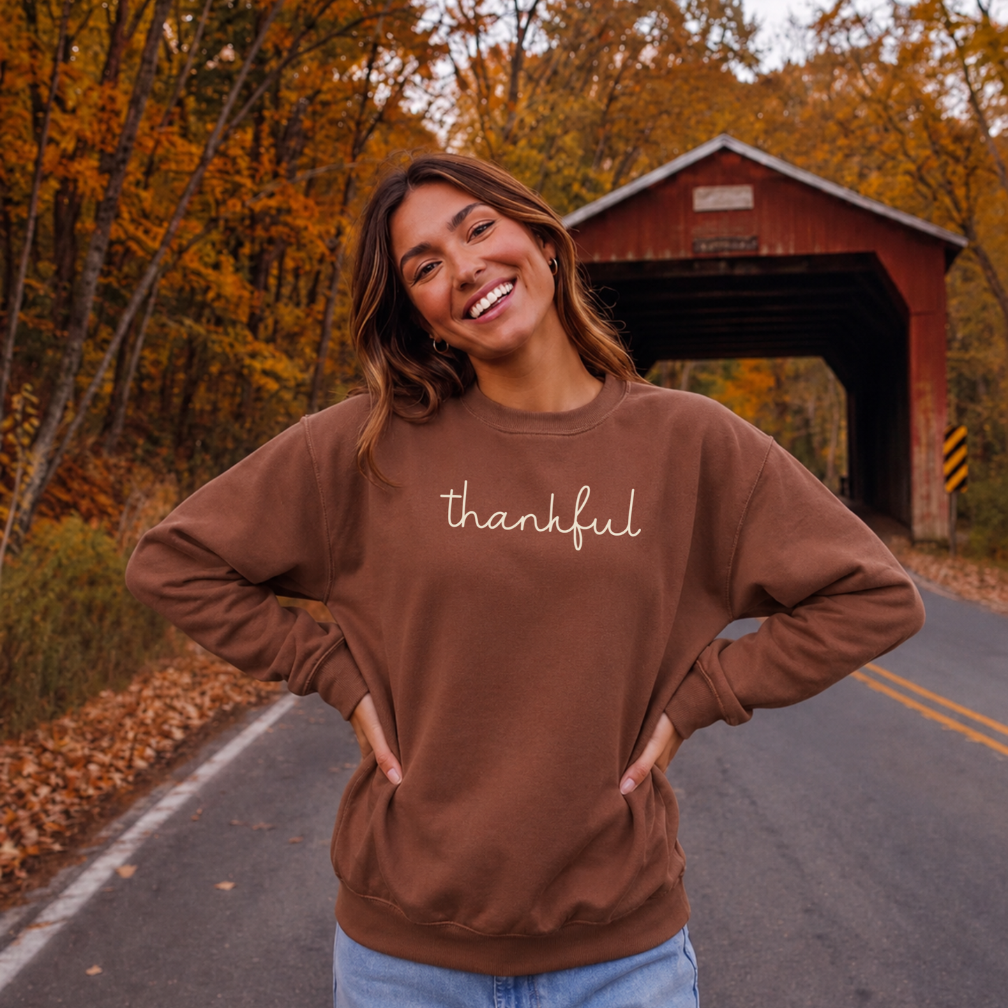Woman wearing brown Thankful script crewneck sweatshirt with cream graphic on a scenic fall road near a covered bridge