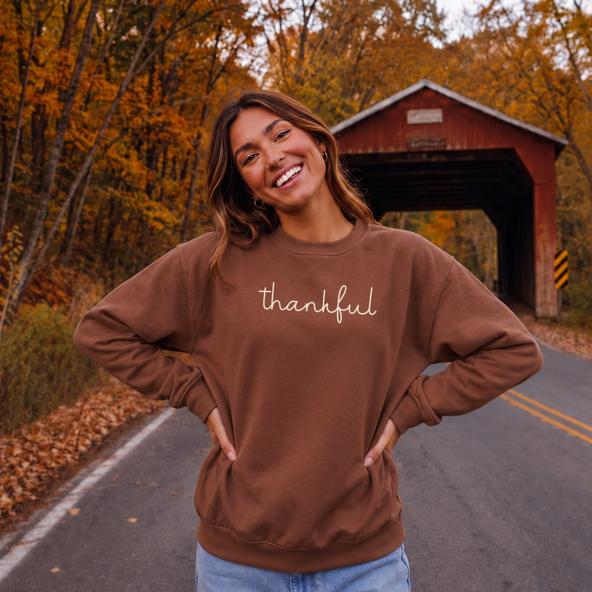 Woman wearing brown Thankful script crewneck sweatshirt with cream graphic on a scenic fall road near a covered bridge