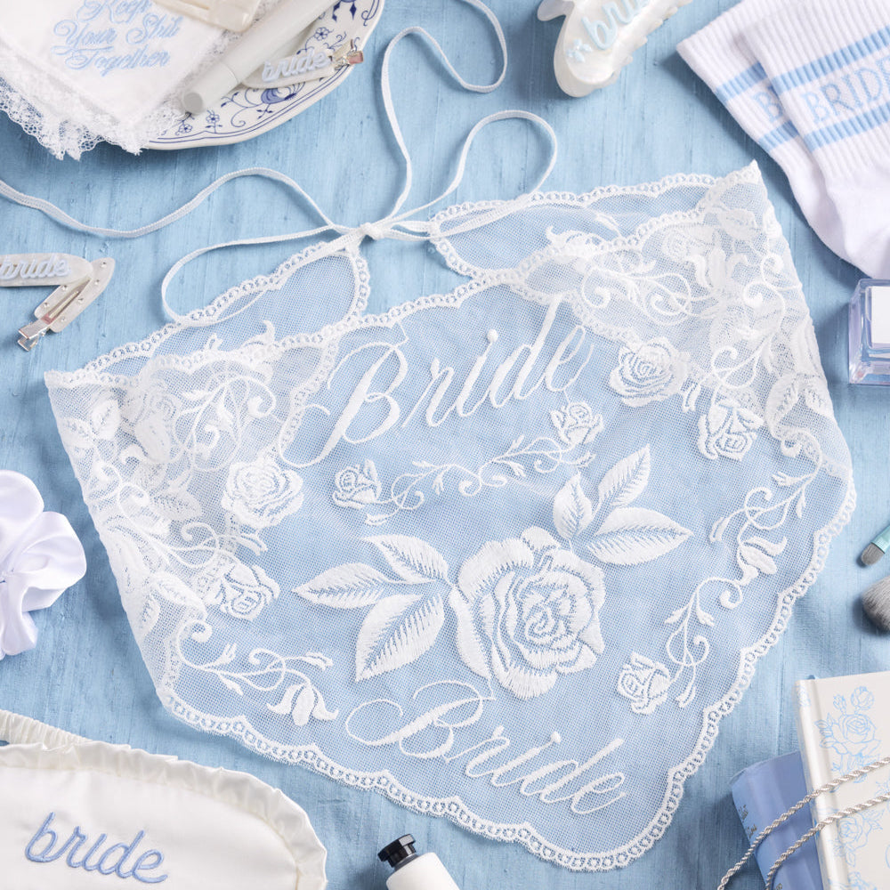 Lace bridal head scarf with 'Bride' embroidery on a blue background with wedding accessories.