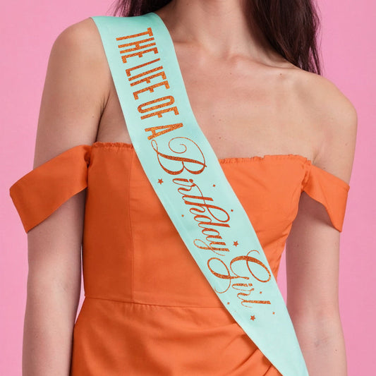 Person wearing an orange dress with a 'Life of a Birthday Girl' sash on a pink background