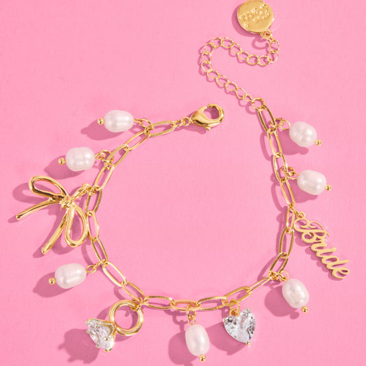 Gold bride charm bracelet featuring bow, rhinestone ring, rhinestone heart and pearl charms on pink background