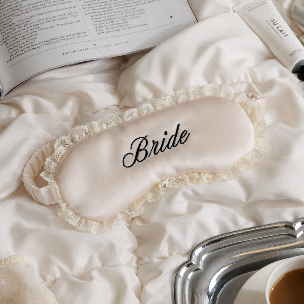 Sleep mask with 'Bride' text on a bed with an open book and coffee cup.