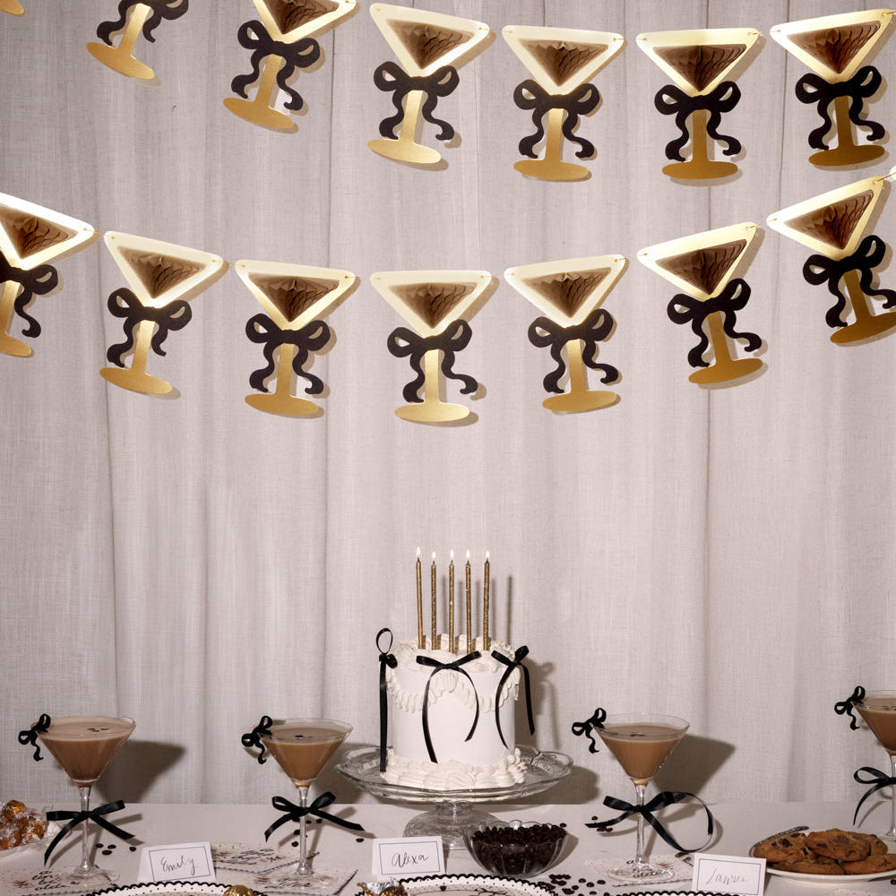 Decorative table setting with a cake, glasses, and a martini-themed banner on a white curtain background.