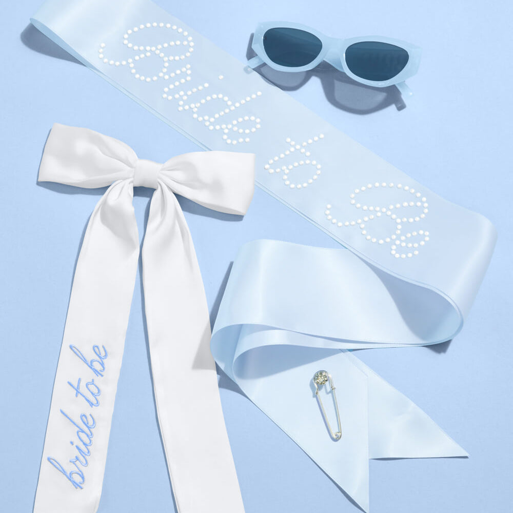 White sash with 'bride to be' text, sunglasses, and a safety pin on a light blue background