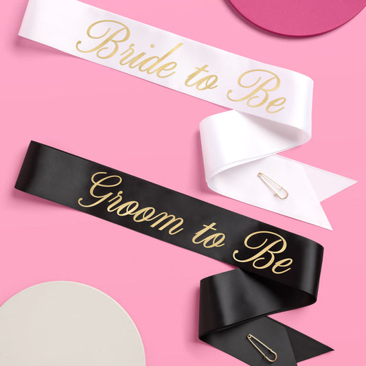 Two sashes, one white with 'Bride to Be' and one black with 'Groom to Be', on a pink background.
