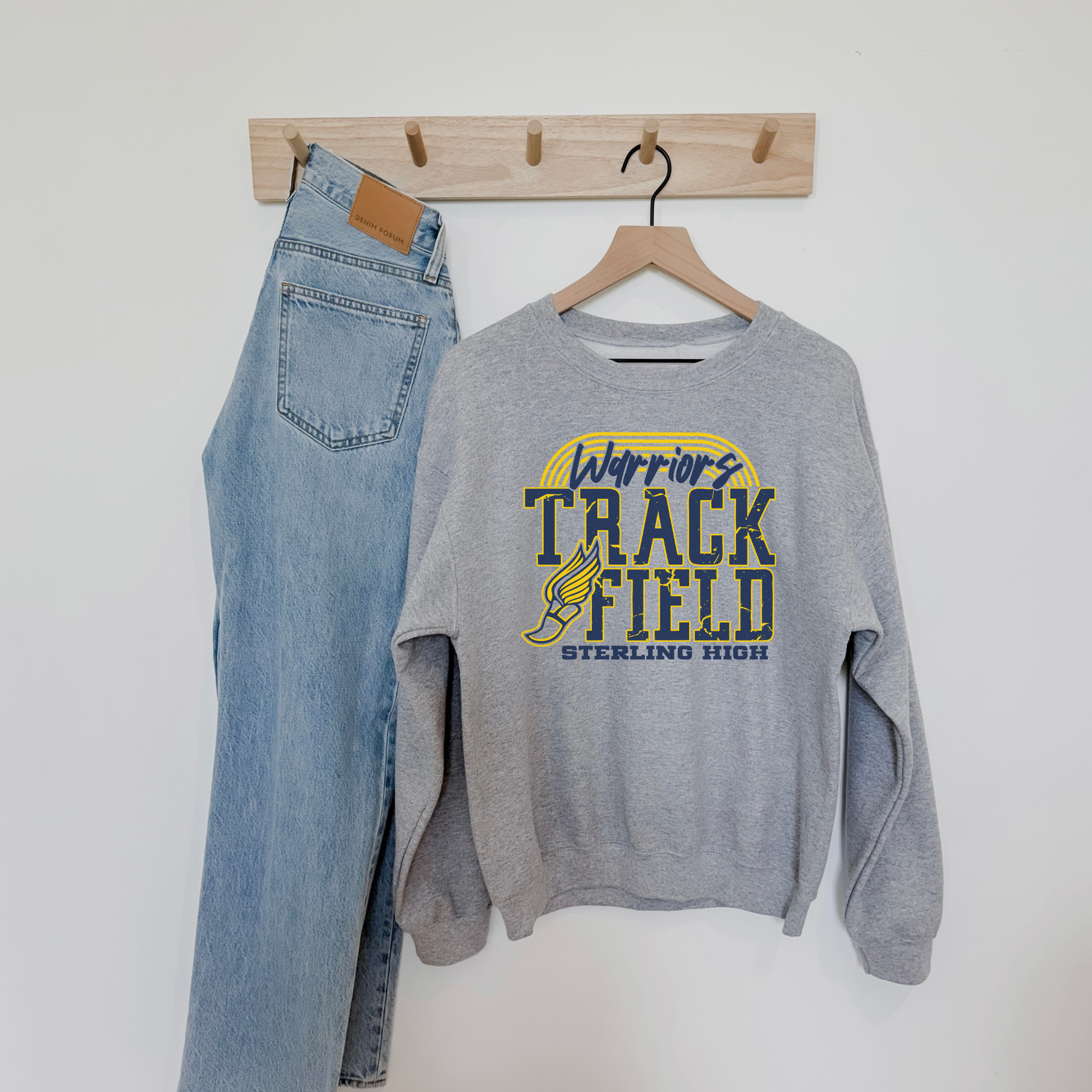 Gray Sterling High Warriors Track and Field sweatshirt styled on a wall hook with denim jeans, showcasing athletic school pride design.