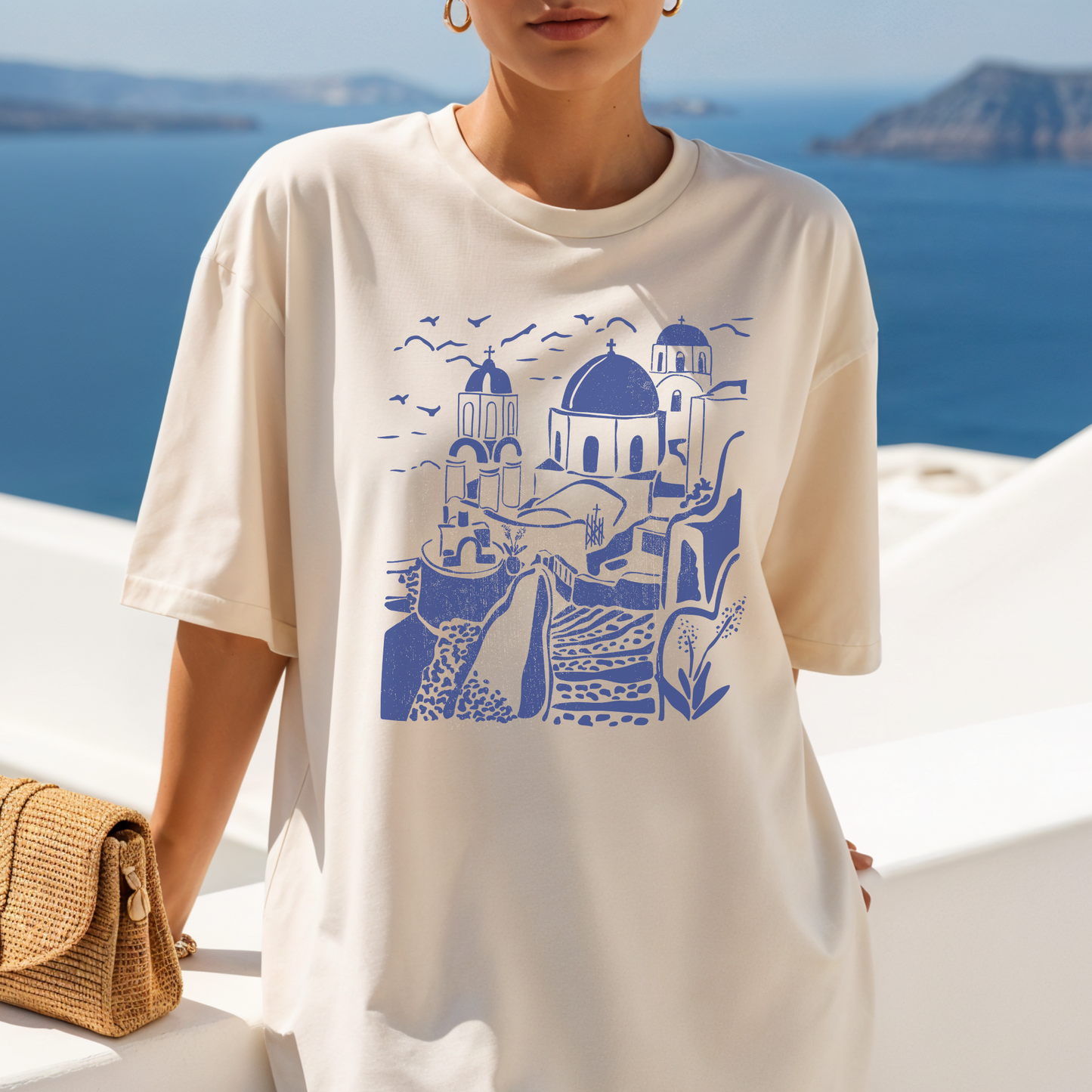 Model wearing a size Large Comfort Colors cream Santorini Greece graphic tee with blue dome illustration, standing on white balcony overlooking ocean.