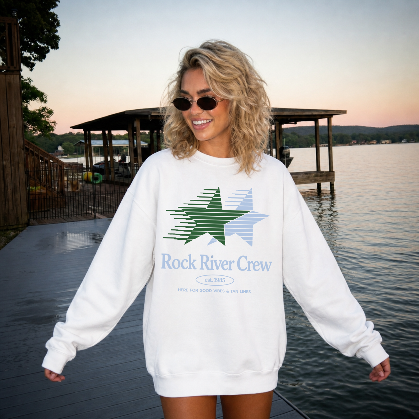Woman wearing a white sweatshirt with a star design and 'Rock River Crew' text by a lake.