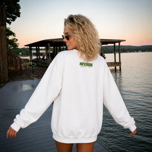 Woman wearing a white sweatshirt with 'HEVYJUDE' branding by a lake at sunset.