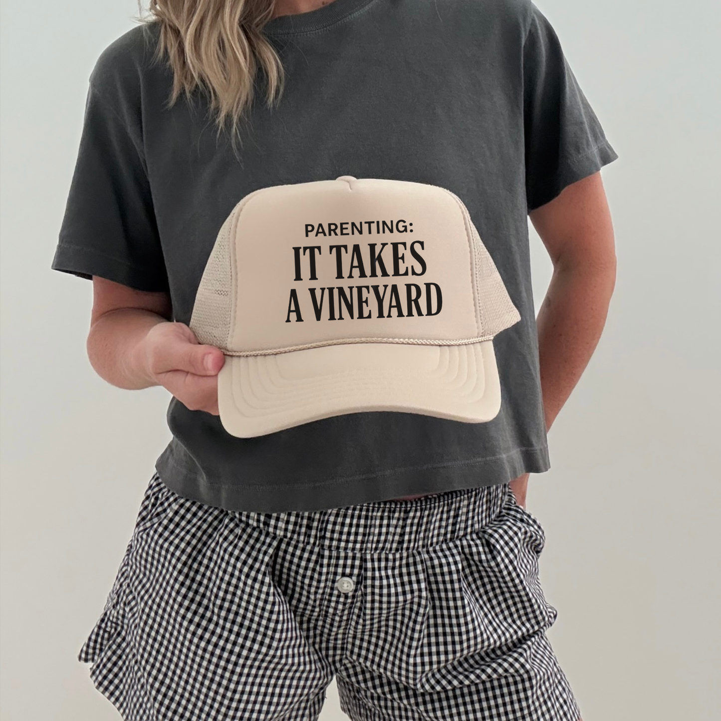 Cream trucker hat with “Parenting: It Takes a Vineyard” text held by model wearing casual outfit