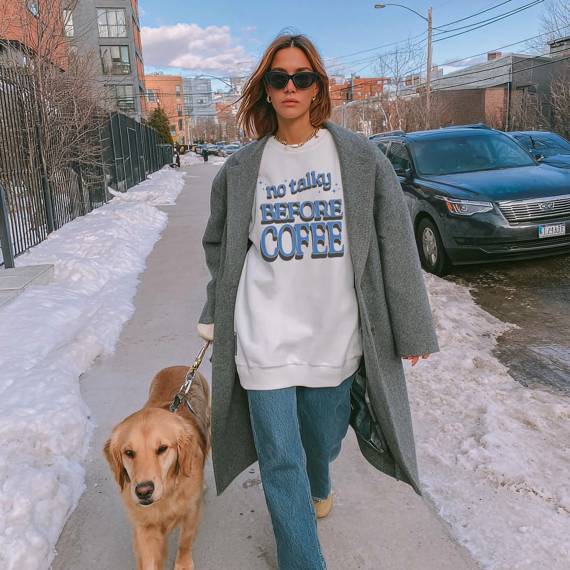 Woman wearing white ‘No Talky Before Coffee’ sweatshirt walking her dog on snowy sidewalk
