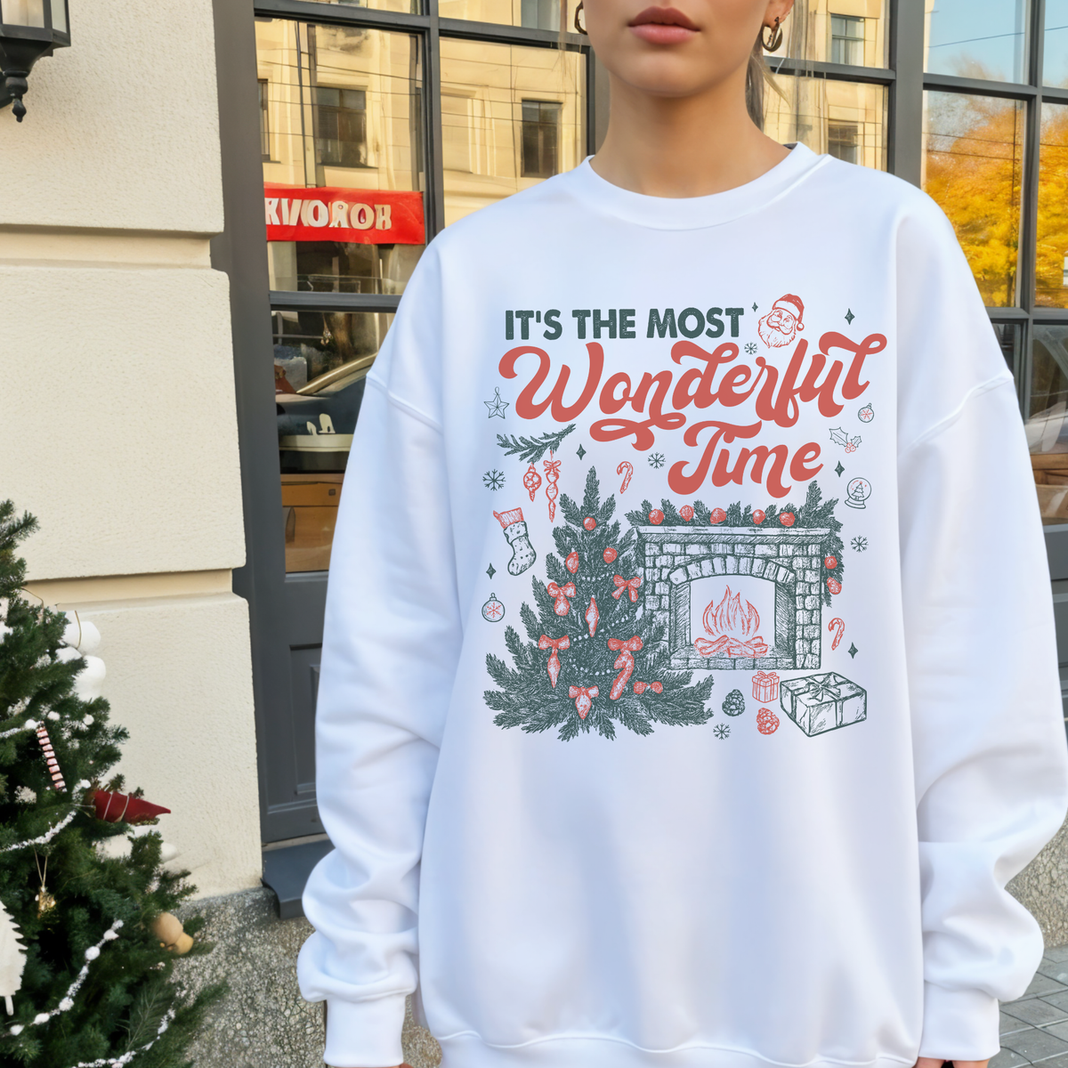 Woman wearing a white “It’s the Most Wonderful Time” Christmas sweatshirt with festive fireplace and tree design, standing near a small Christmas tree outside a shop.