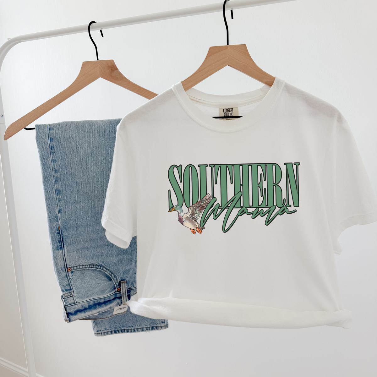 White Southern Mama tee hanging next to denim jeans, featuring green lettering and duck illustration.