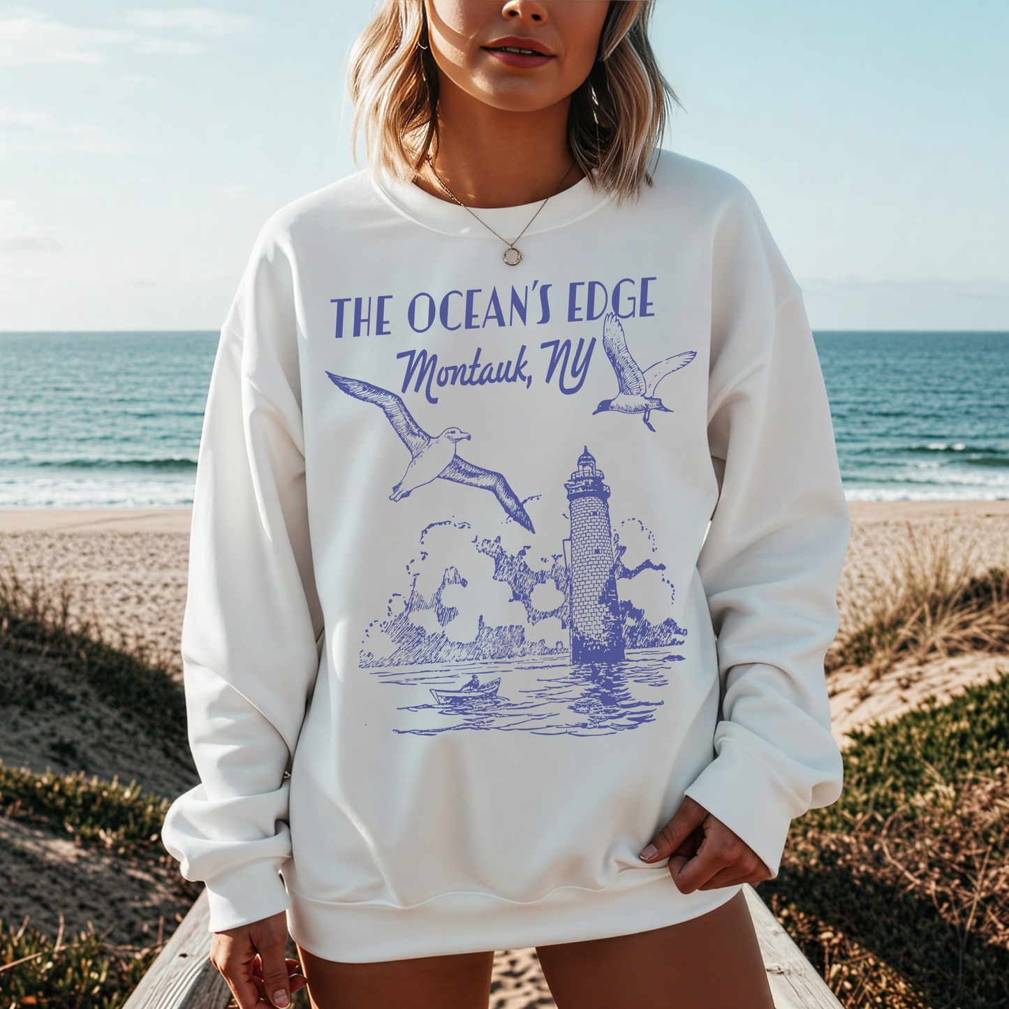 "Woman wearing white Montauk lighthouse graphic sweatshirt standing by the ocean."