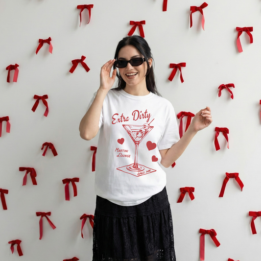 Model wearing white Extra Dirty Martini graphic tee in front of red ribbon wall, smiling with sunglasses.