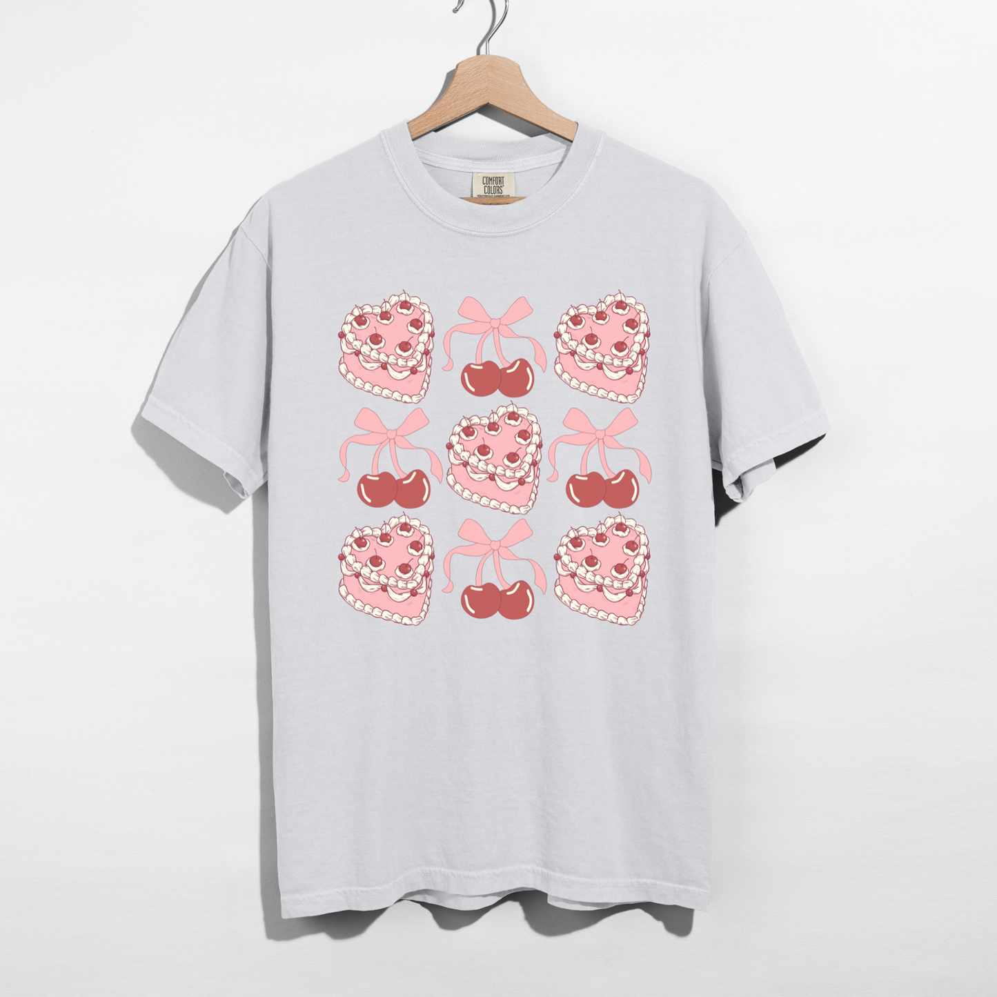 Sweetheart Cherry Cake Graphic Tee with pink heart cakes, cherries, and bows on a white shirt hanging on a wooden hanger.