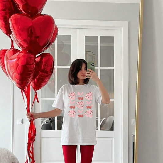 Woman wearing a white Sweetheart Cherry Cake Graphic Tee holding red heart balloons in a mirror selfie.