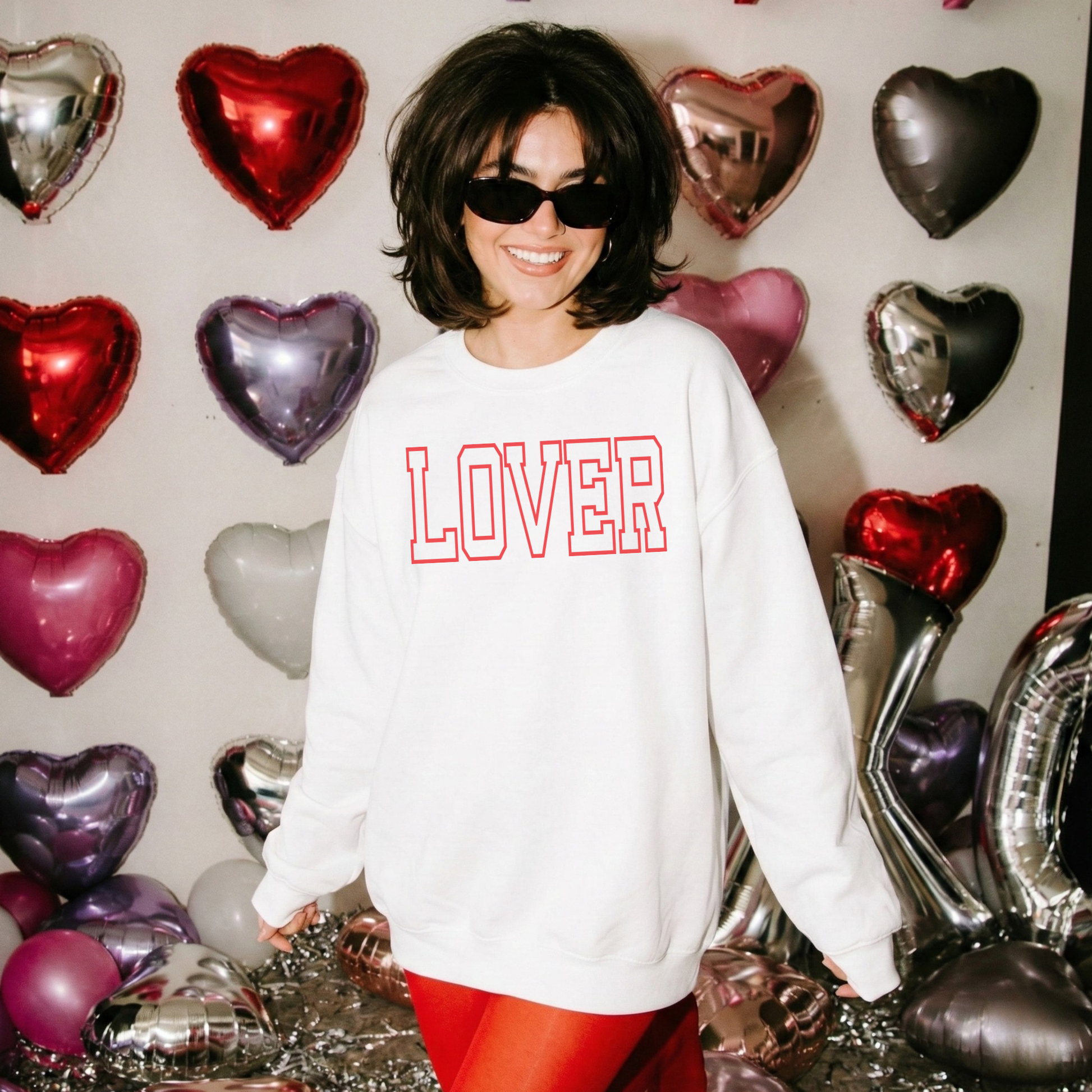 “Woman wearing white LOVER sweatshirt with red varsity lettering, standing in front of heart balloon backdrop.”