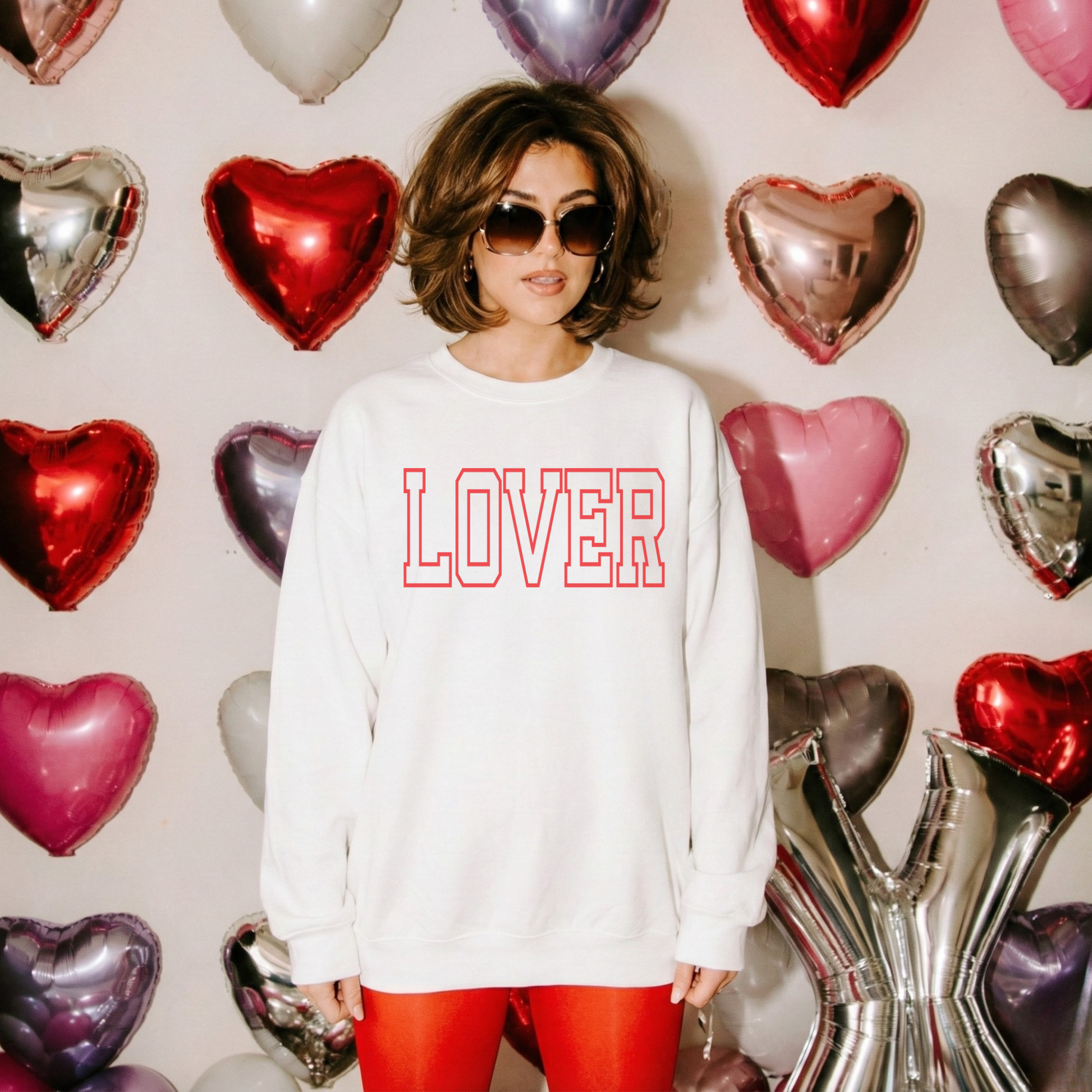 Woman in white LOVER sweatshirt with red lettering and oversized sunglasses in a Valentine balloon backdrop.