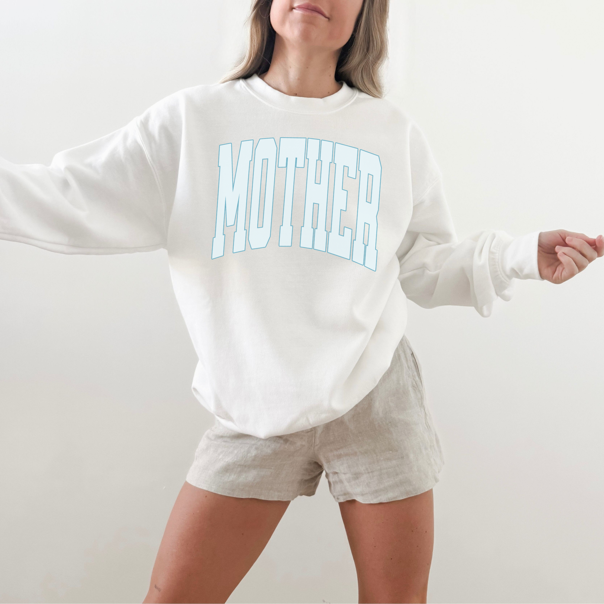 Woman wearing white MOTHER collegiate sweatshirt styled with neutral shorts in bright studio setting
