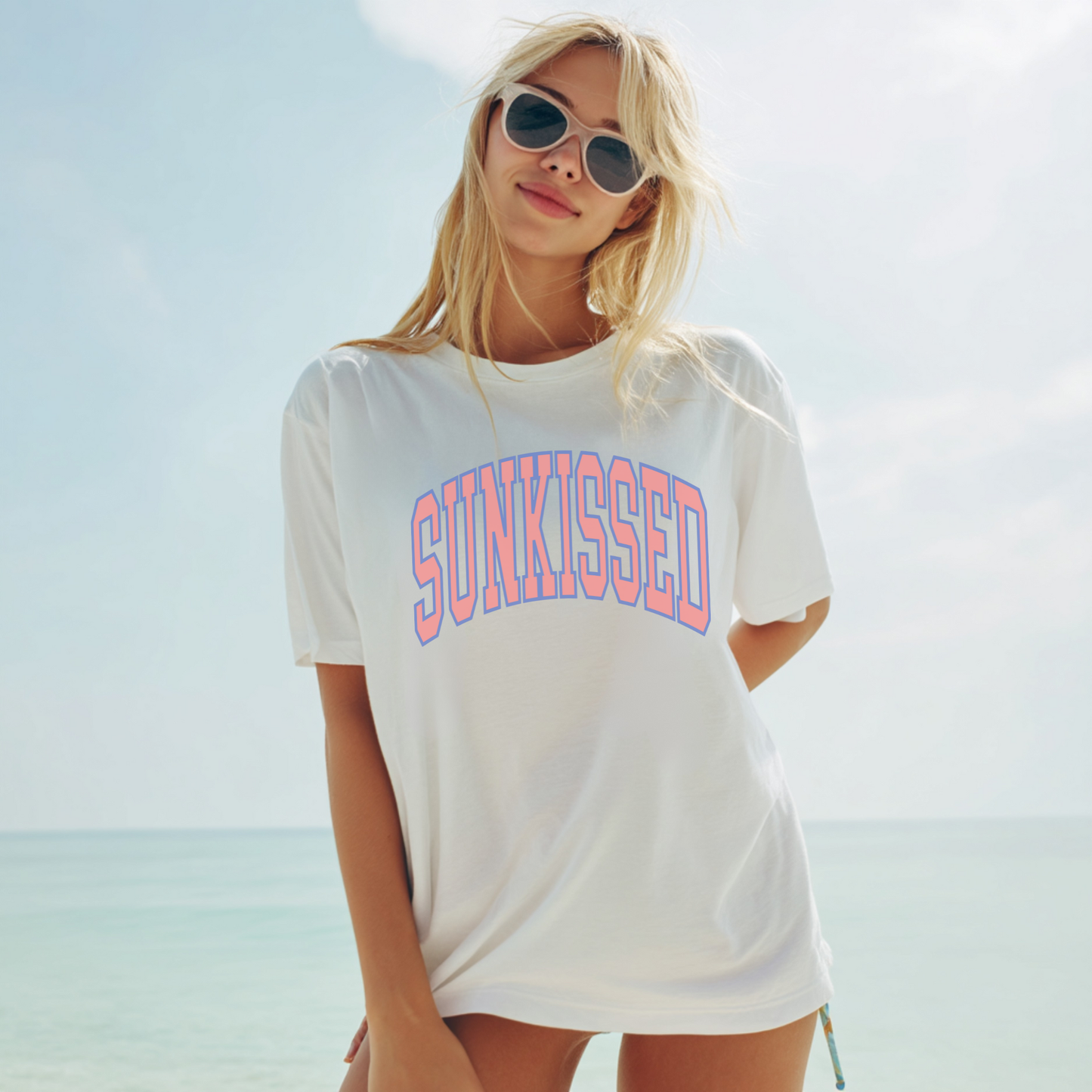 Woman wearing a white oversized SUNKISSED graphic tee styled as a beach cover-up, relaxed summer outfit by the ocean