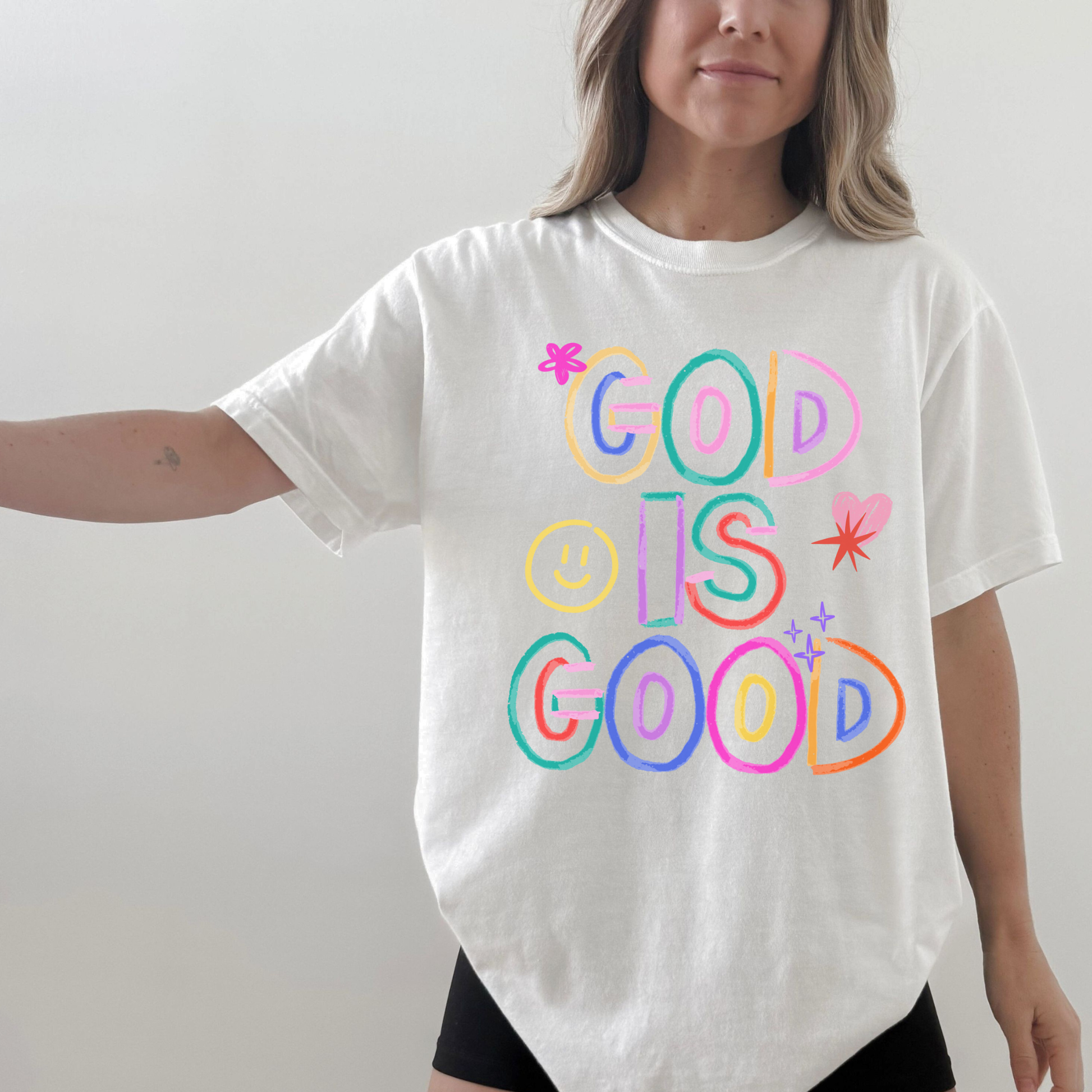 Person wearing a white t-shirt with 'GOD IS GOOD' in colorful letters on a plain background