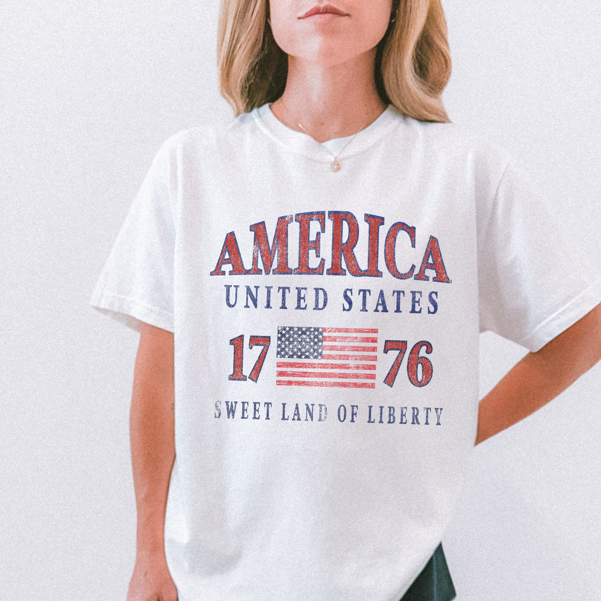 Person wearing a white t-shirt with 'America United States 1776 Sweet Land of Liberty' text and American flag design.