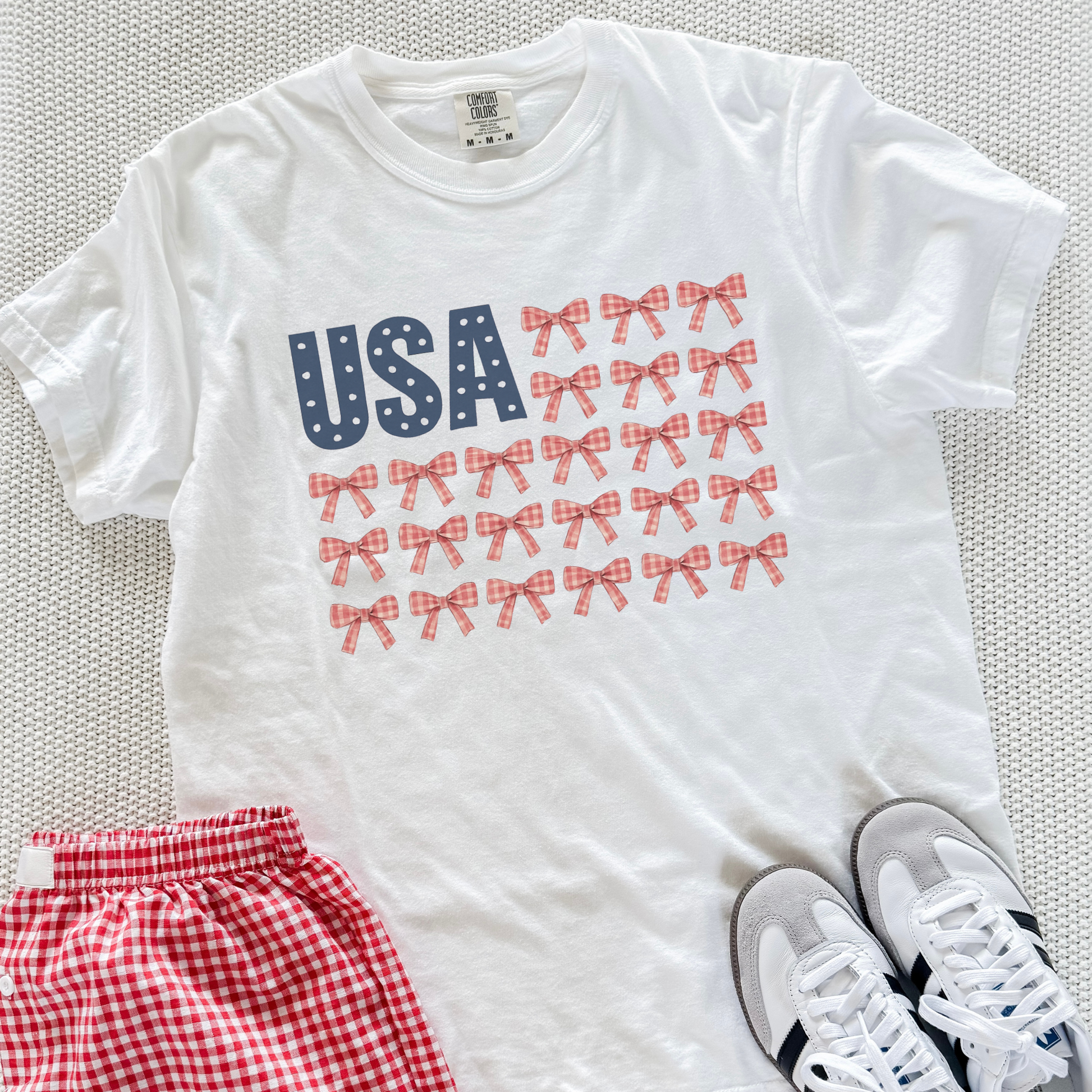 White t-shirt with 'USA' and bow design, paired with red checkered shorts and white sneakers on a textured surface.