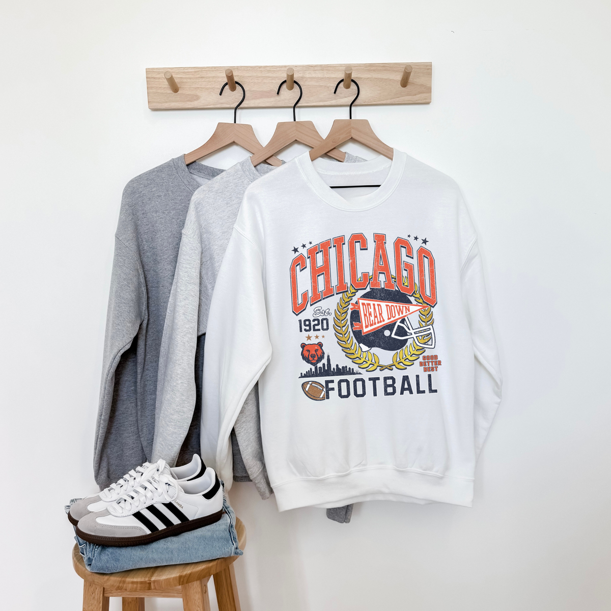 White sweatshirt with 'Chicago Football' design on a wooden rack with gray sweater and sneakers.