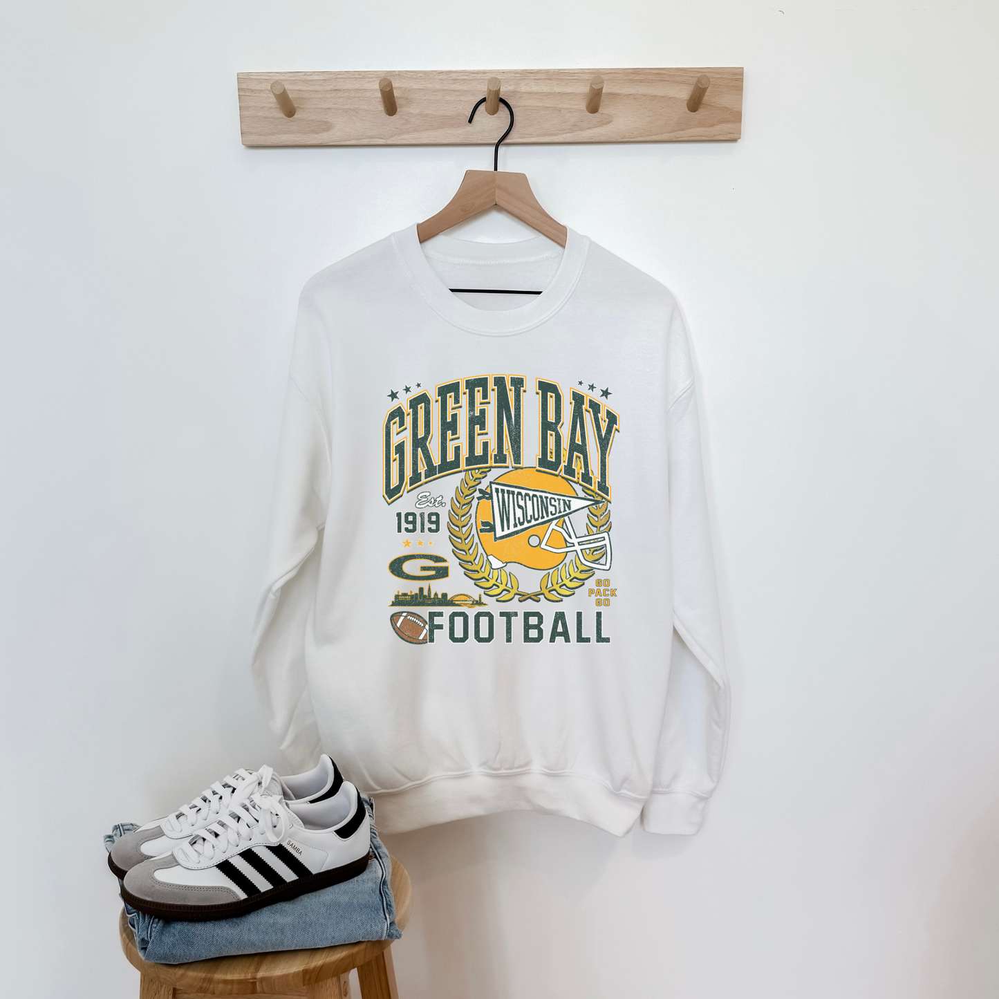 White sweatshirt with 'Green Bay Football' design on a wooden stool against a white wall.
