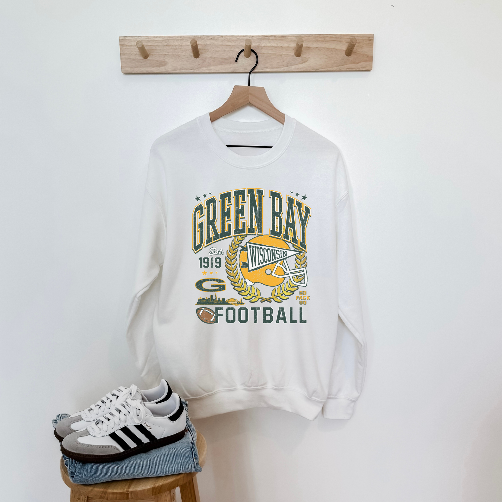 White sweatshirt with 'Green Bay Football' design on a wooden stool against a white wall.