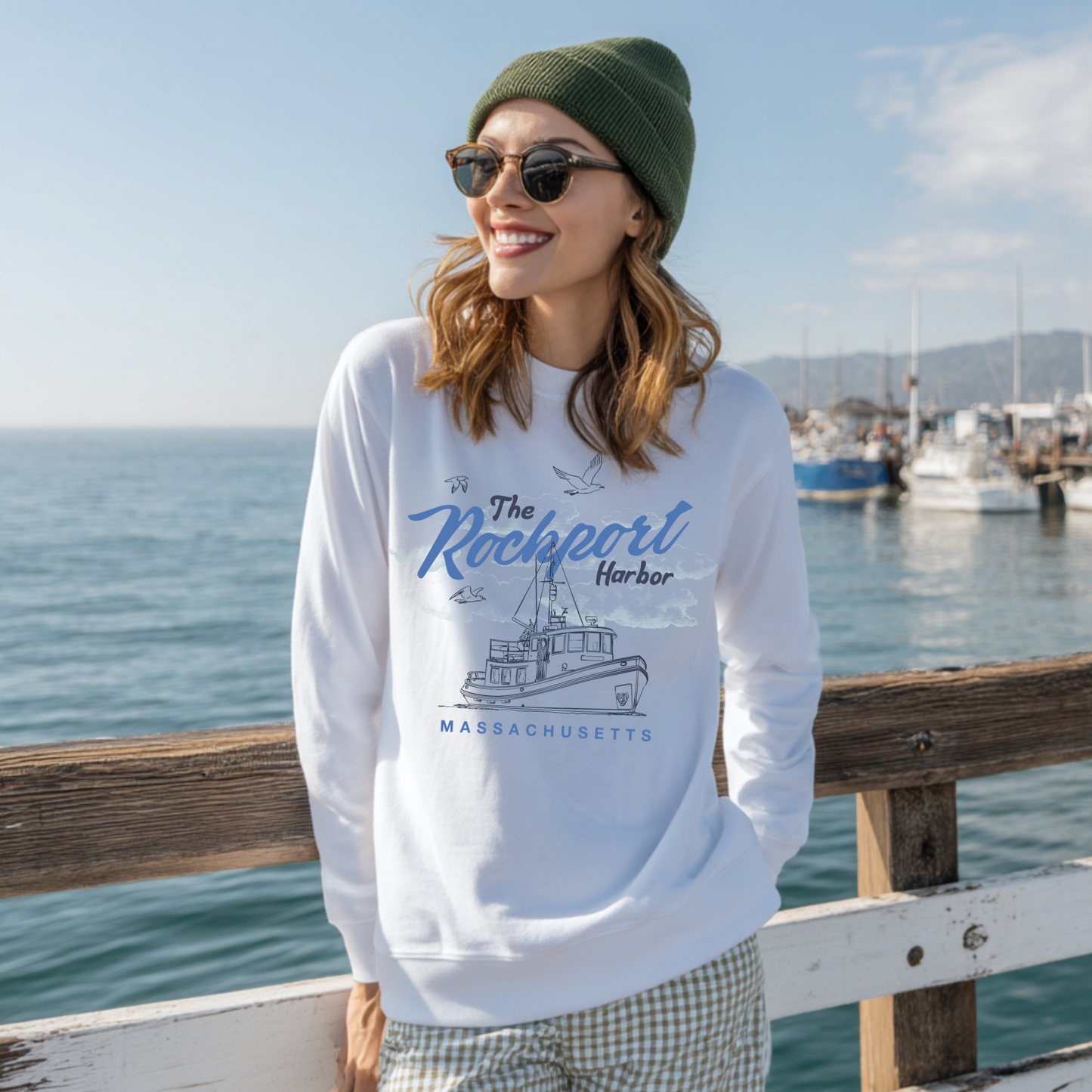 Woman wearing white Rockport Harbor Massachusetts crewneck sweatshirt standing by ocean pier with fishing boats in background