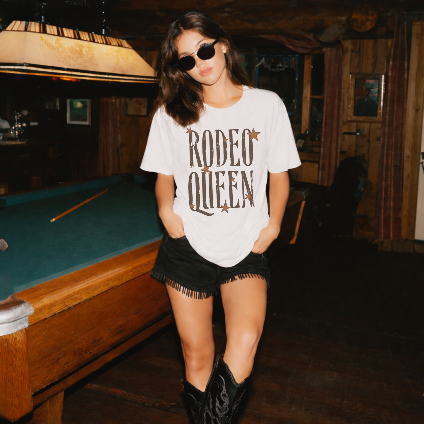 Model wearing Rodeo Queen graphic t-shirt with black shorts and cowboy boots in rustic western bar