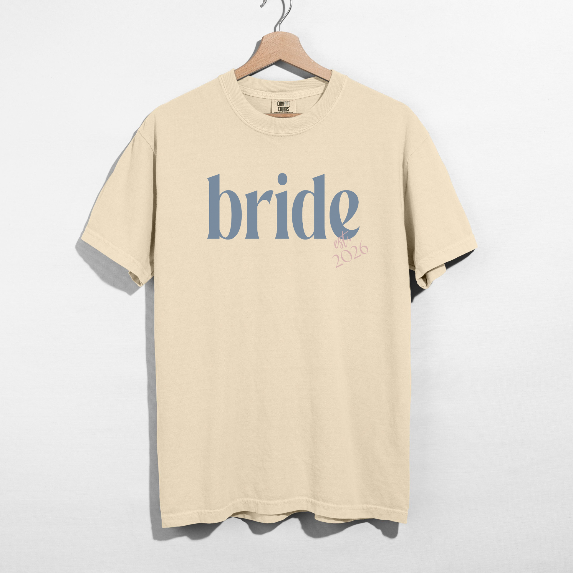 Beige t-shirt with 'bride' printed on it, hanging on a wooden hanger against a white background.