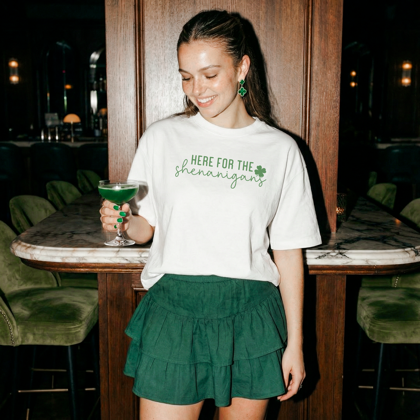 Woman wearing white Here for the Shenanigans shirt styled with green skirt and clover earrings for St Patrick’s Day