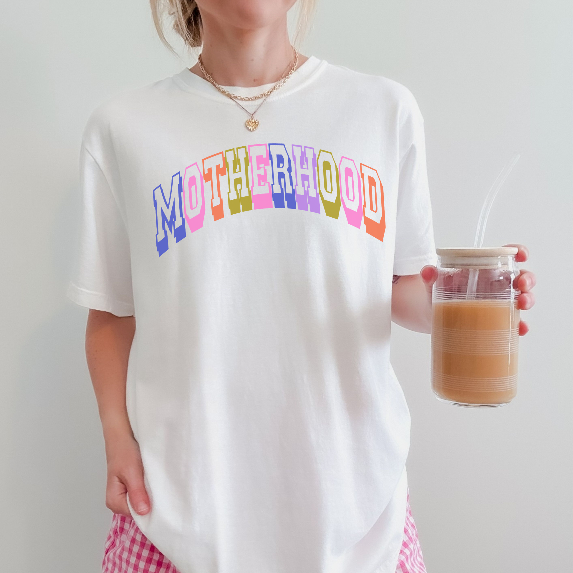 Woman wearing a white Comfort Colors 1717 graphic tee featuring colorful retro collegiate “Motherhood” lettering while holding iced coffee.