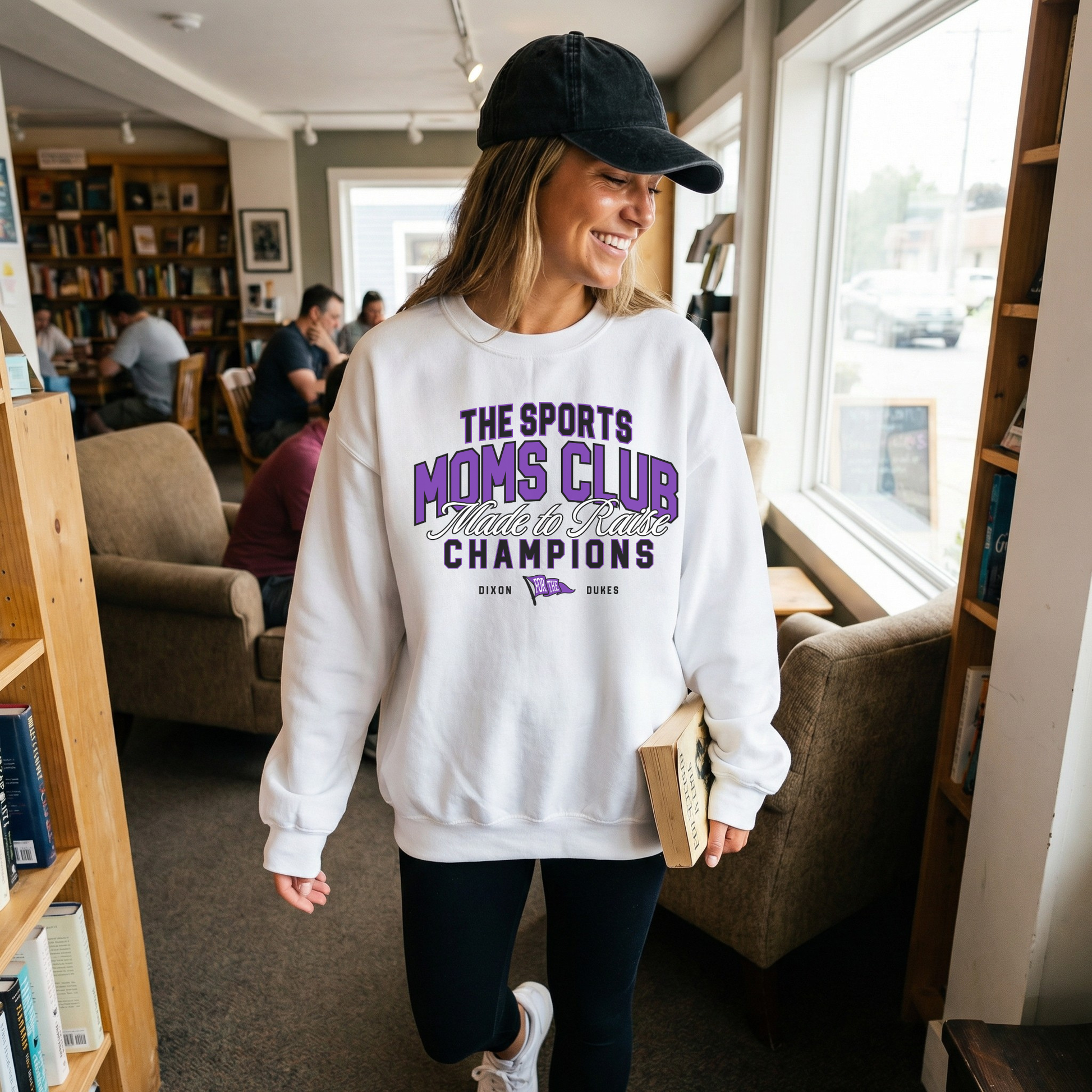 White Sports Moms Club sweatshirt worn in cozy coffee shop setting smiling lifestyle photo
