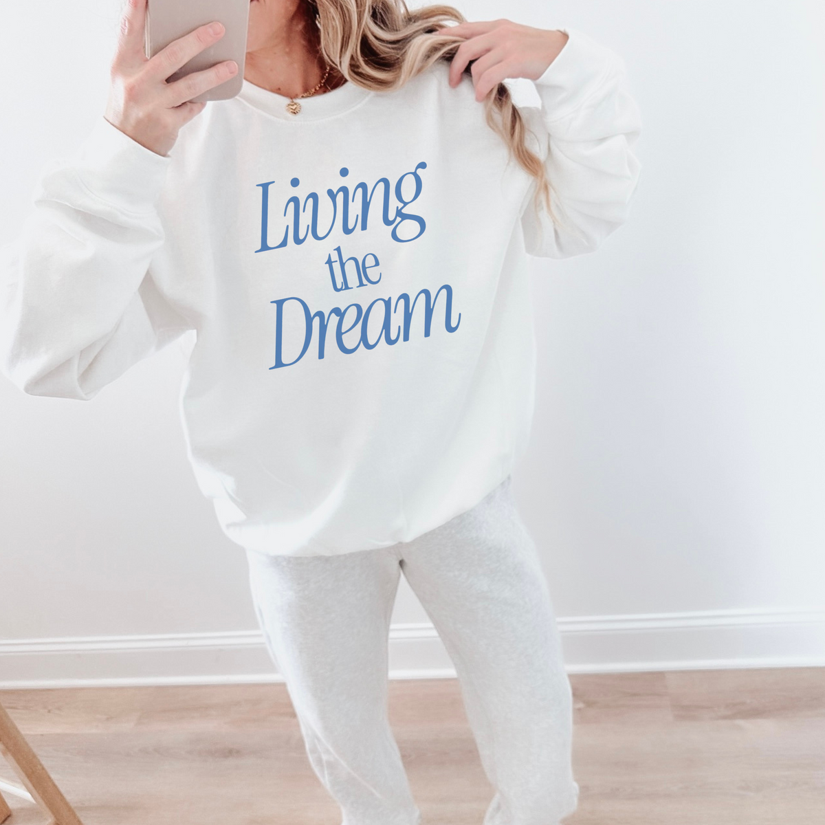 Person wearing a white sweatshirt with 'Living the Dream' text in blue, holding a phone.