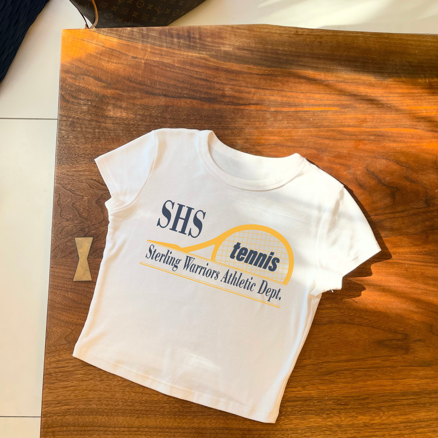 White cropped women’s tennis shirt featuring SHS Sterling Warriors Athletic Department tennis graphic.
