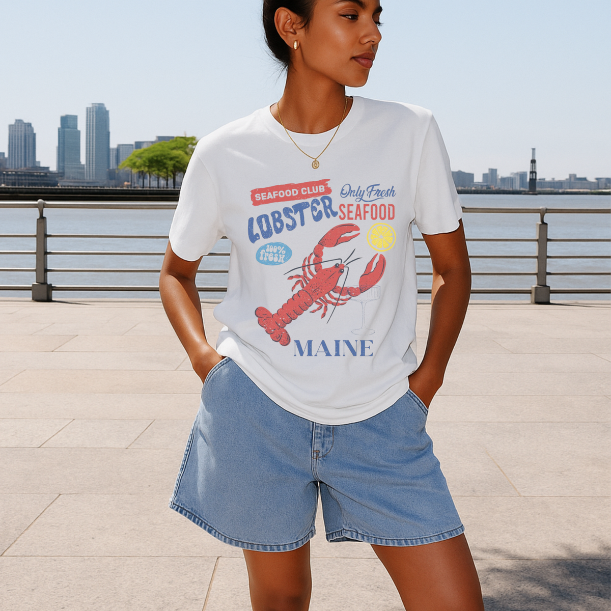 Woman wearing a white t-shirt with a lobster graphic and 'Maine' text, standing by a waterfront with city skyline in the background.