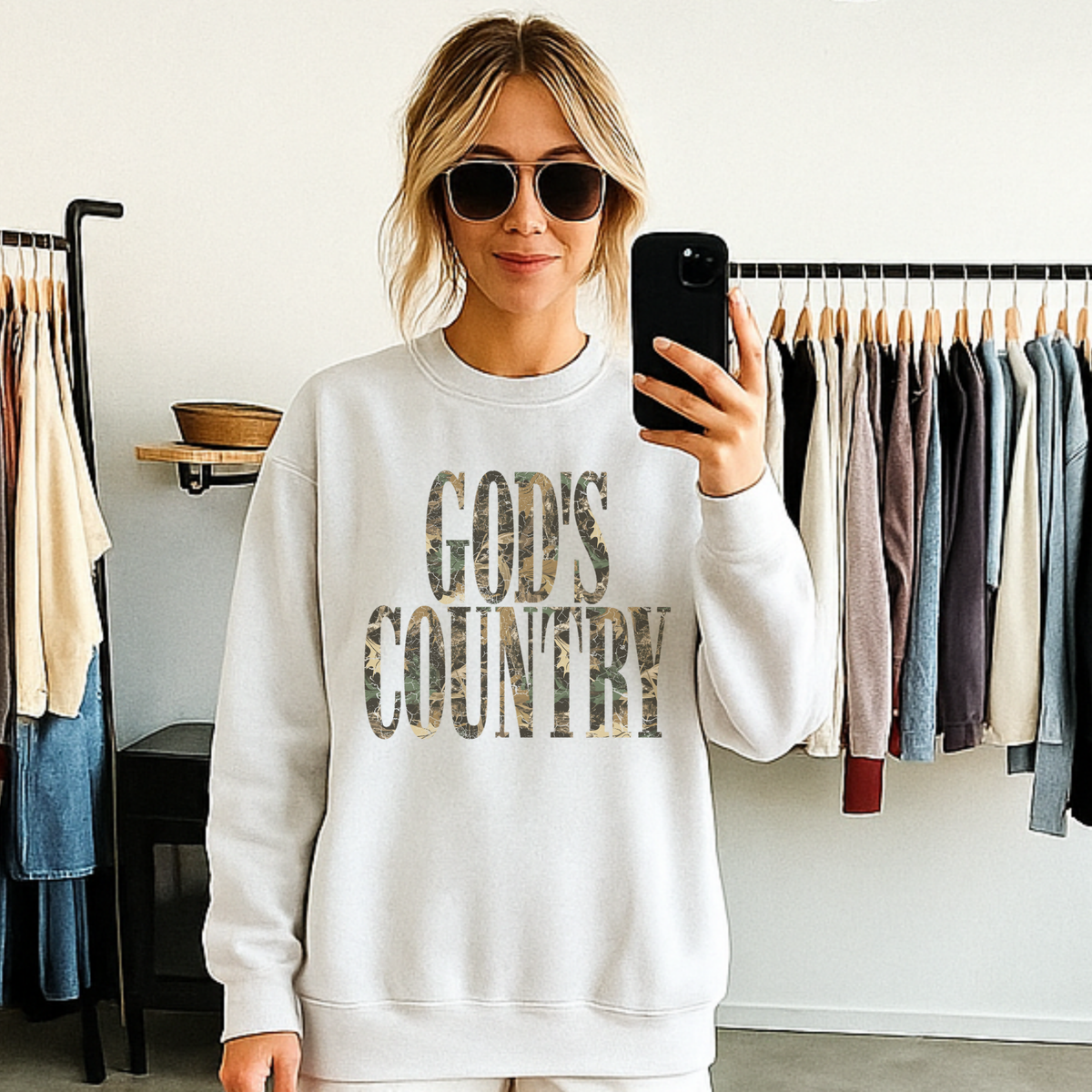 Woman wearing God’s Country camo graphic crewneck sweatshirt styled with sunglasses in a boutique setting