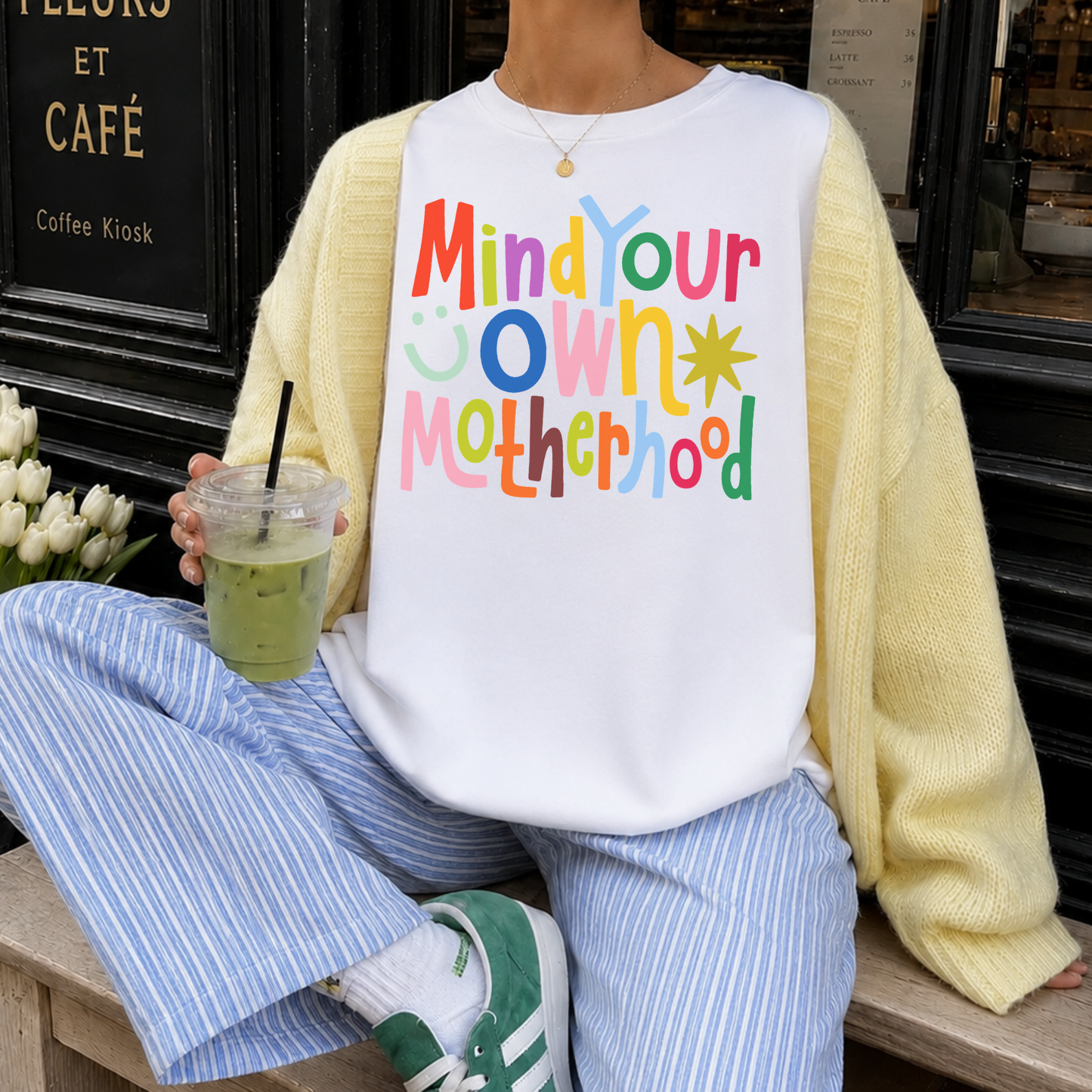 Woman wearing colorful Mind Your Own Motherhood graphic tee styled with yellow cardigan and striped pants holding iced matcha latte outside cafe