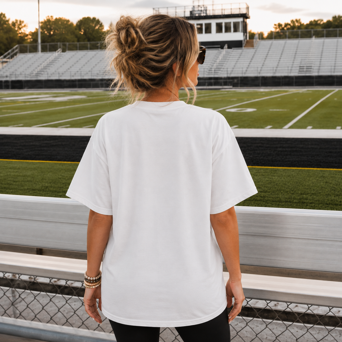 Back view of woman wearing white football game day graphic tee at stadium bleachers