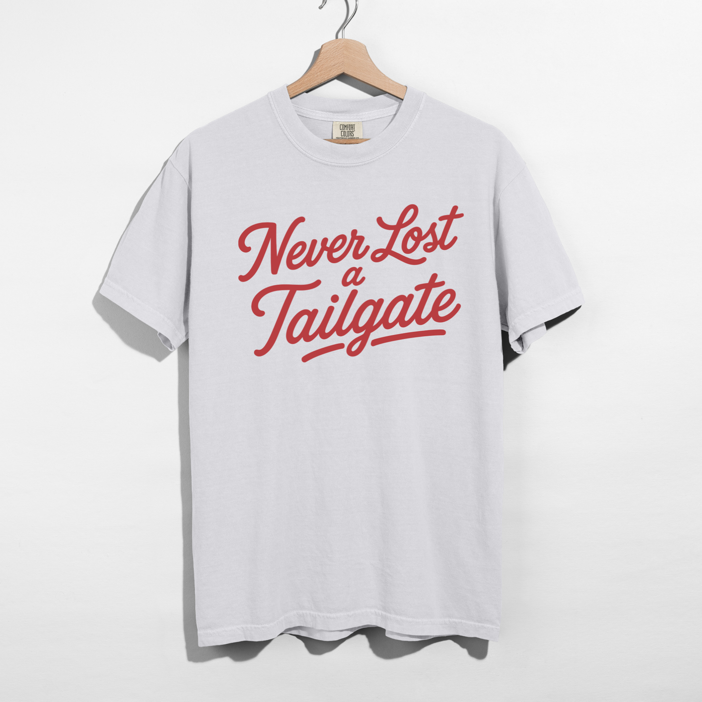 White graphic T-shirt featuring the phrase "Never Lost a Tailgate" in crimson vintage script lettering hanging on a wooden hanger.
