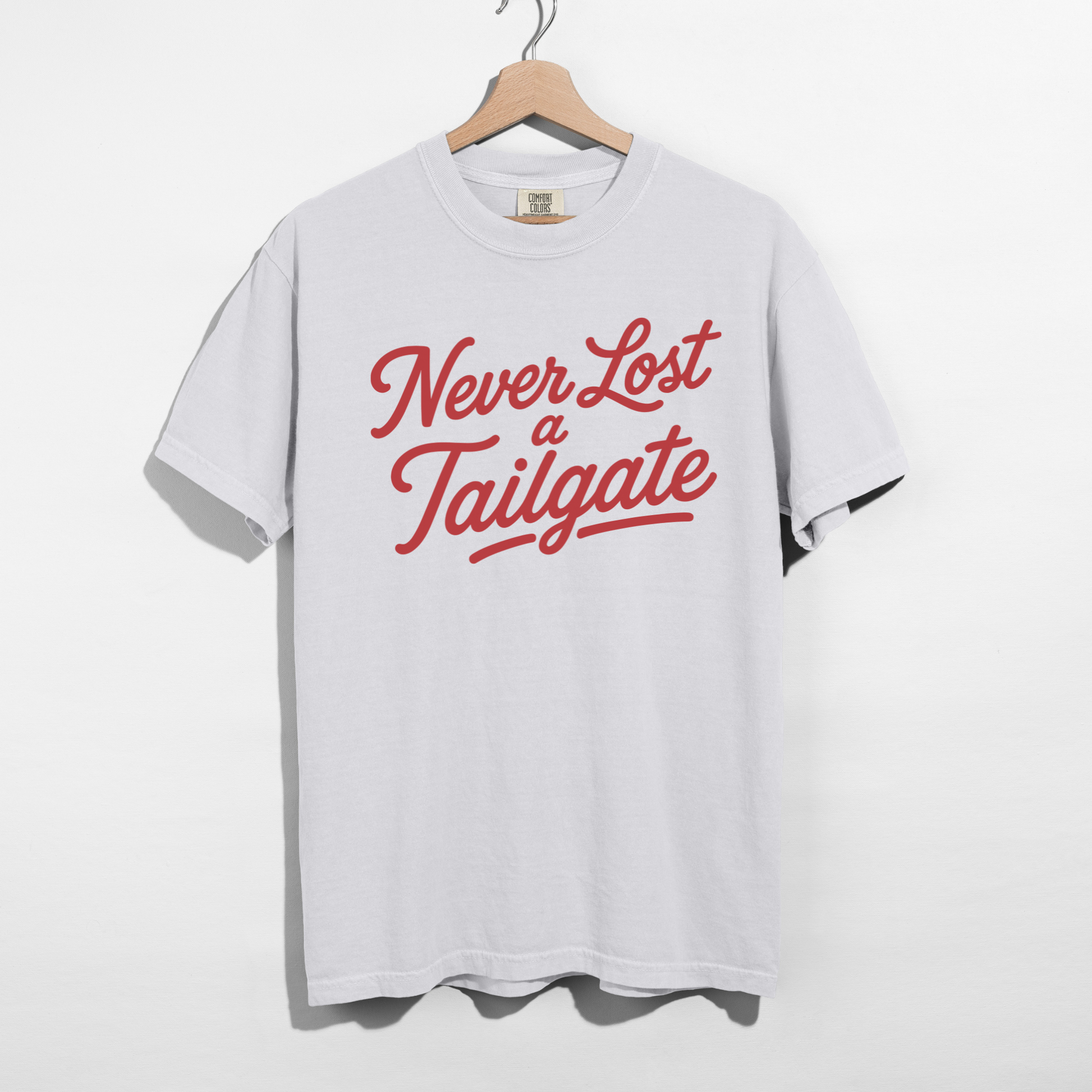 White graphic T-shirt featuring the phrase "Never Lost a Tailgate" in crimson vintage script lettering hanging on a wooden hanger.