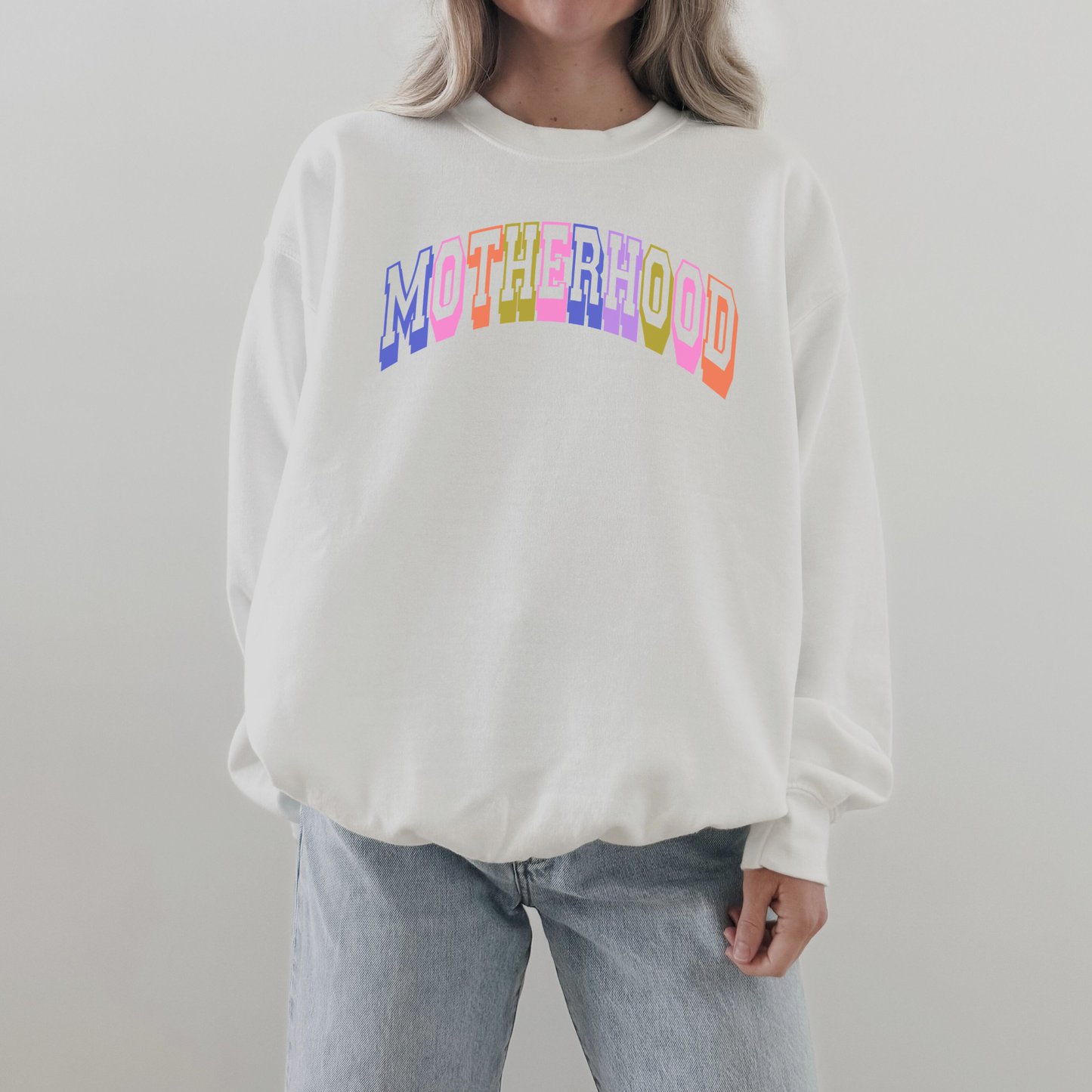 Woman wearing oversized white retro motherhood crewneck sweatshirt with colorful collegiate lettering and light wash jeans.