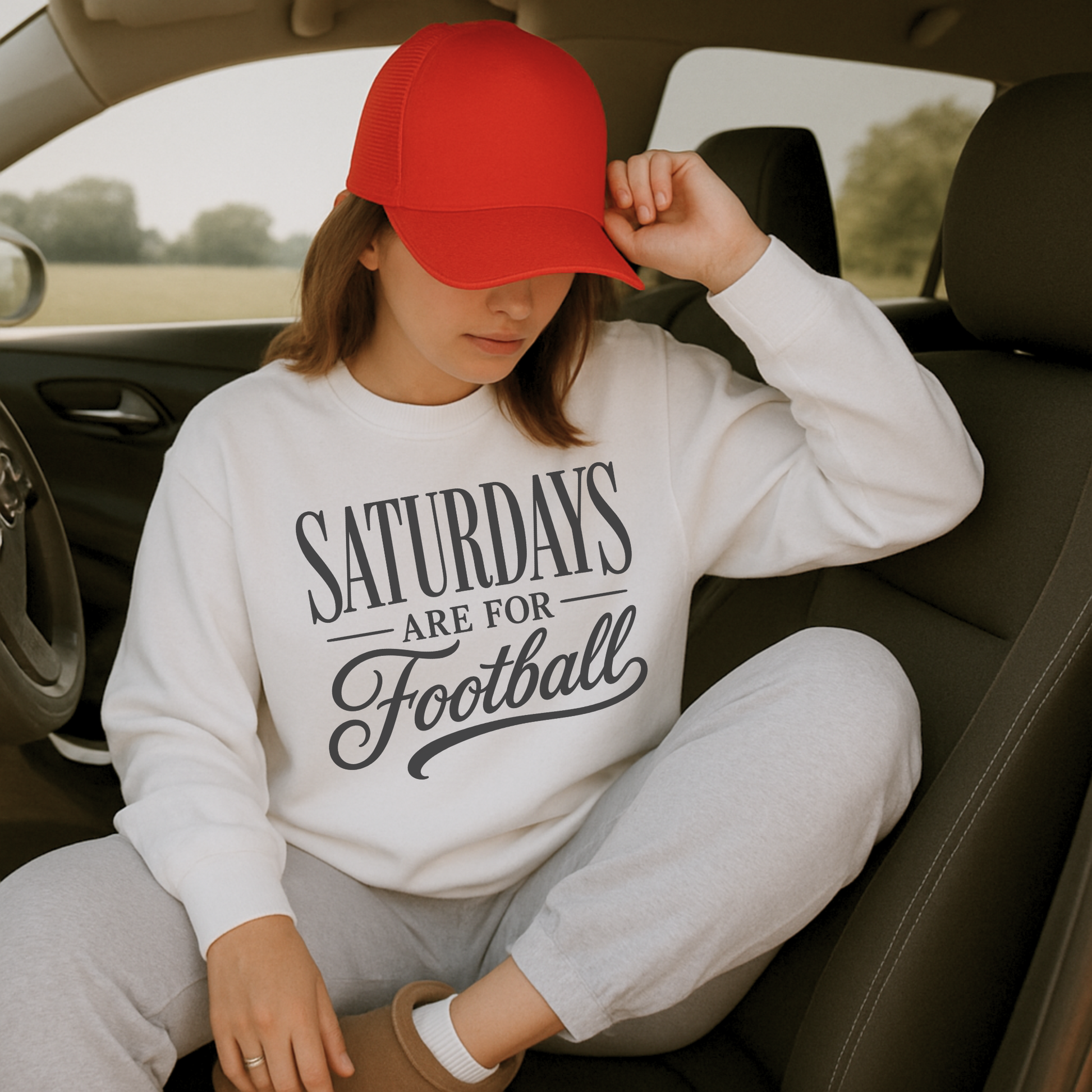 Woman wearing a white "Saturdays Are For Football" graphic sweatshirt with gray joggers and a red trucker hat while sitting inside a vehicle before game day.