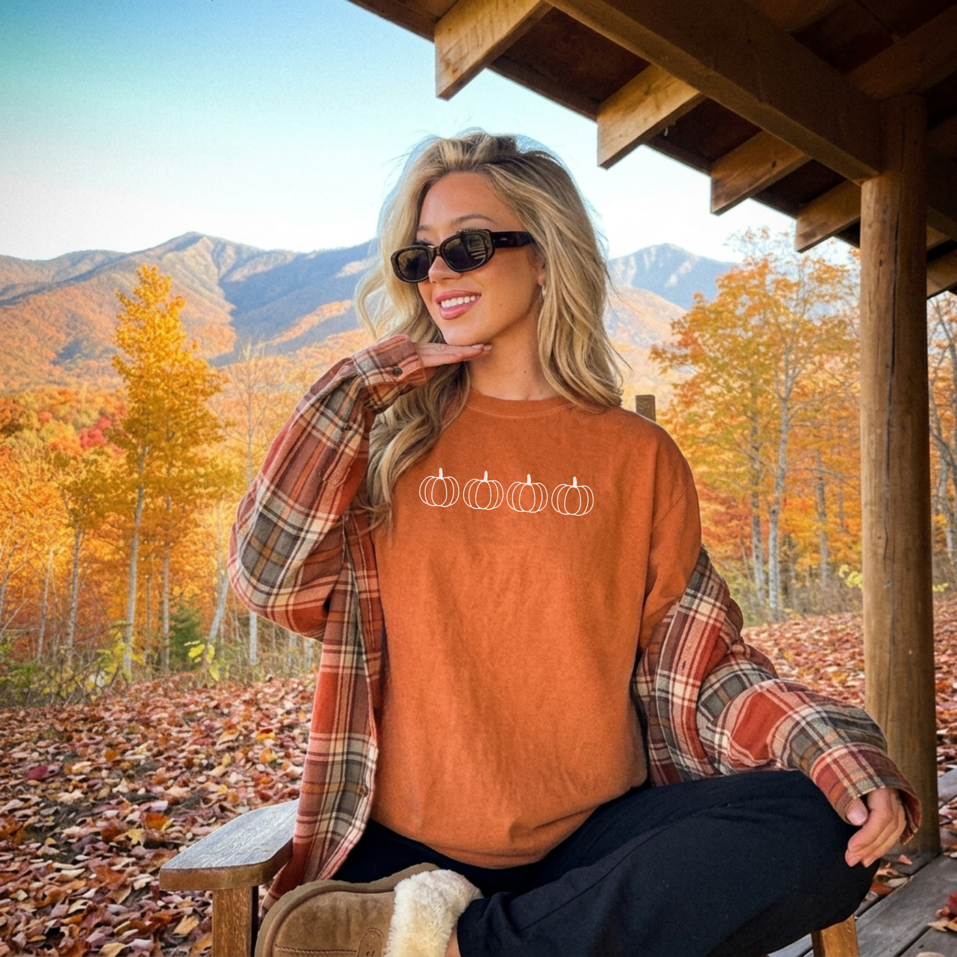 Woman wearing a rust orange minimalist pumpkin graphic tee with four white outline pumpkins layered under a plaid flannel in a beautiful mountain fall landscape.