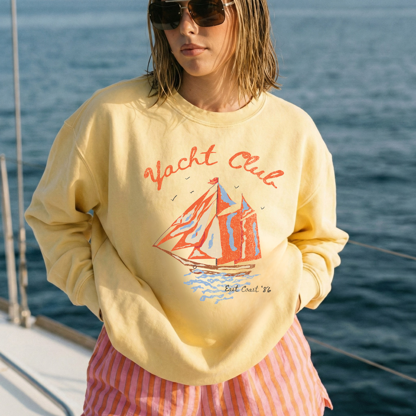 woman wearing yellow yacht club graphic sweatshirt on boat coastal summer outfit sunset water